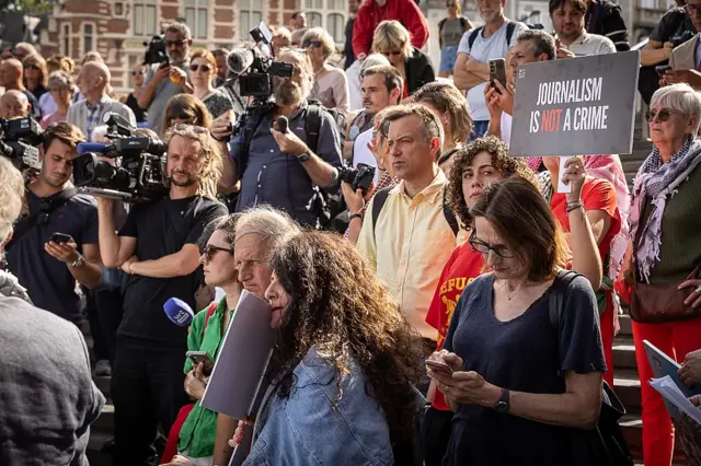 A crowd of people gather at a protest in Brussels. One of them is holding a black placard with red and white writing which reads "Journalism is not a crime" in capitals. Other people are holding TV cameras on their shoulders.