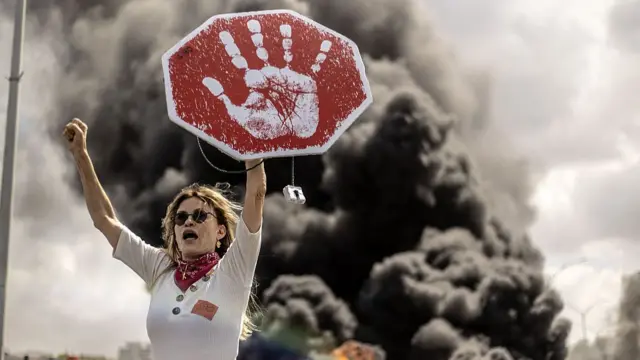 A woman holds her arm up as black smoke billows towards the sky behind her