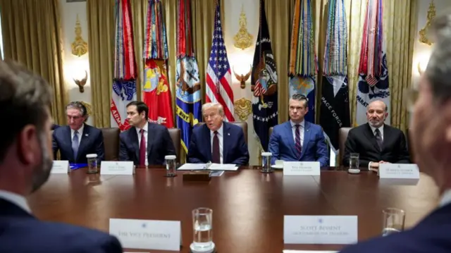 Trump and administration sit behind a dark brown table, behind them is a golden eagle - beneath and above it is gold trim.