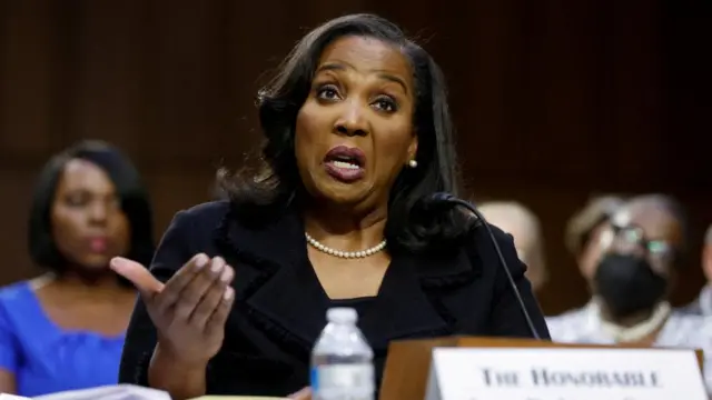 Lisa Cook testifies before a Senate Banking Committee hearing on her nomination to be a member of the Federal Reserve Board of Governors. She gestures with her hand and wears and all black suit with a white pearl neckplace.