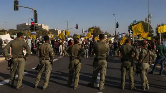 A row of soldiers in khaki military fatigues faces off on a road against protestors holding yellow banners
