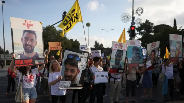 A group of protestors holding photos of hostages and yellow flags in a street at twlight