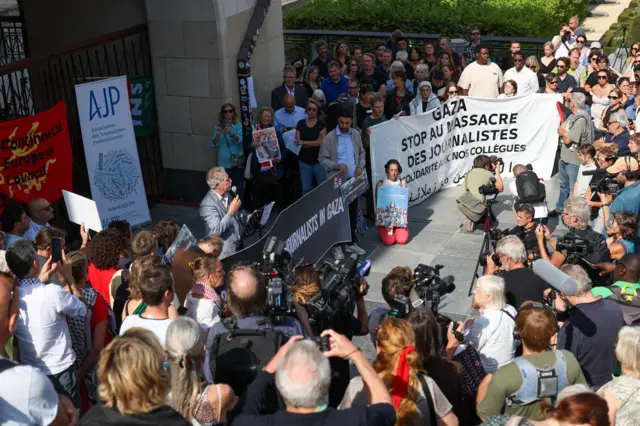 (A large gathering of people at a protest in Brussels. In the centre of the crowd a man is speaking holding a microphone. A woman is kneeling down on the stone floor holding a picture of the journalists who have died reorting in Gaza. People either side of her hold banners reading "Gaza stop au massacre des journalistes".