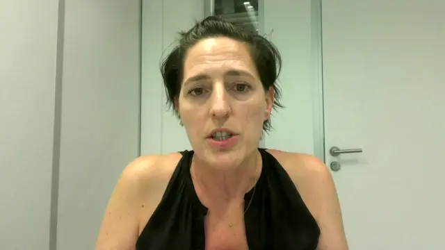 A woman with short black hair and a black vest top speaks from a white room