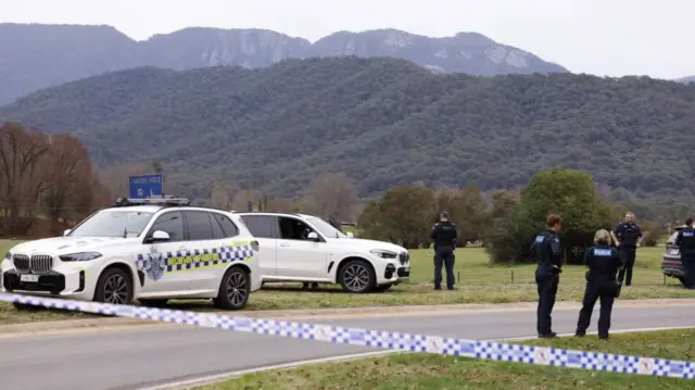 Police cars are seen behind police tape, with four officers and rolling hills in the background