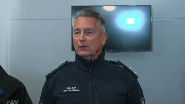 Mike Bush, Victoria Police Commissioner stood in uniform, TV behind him