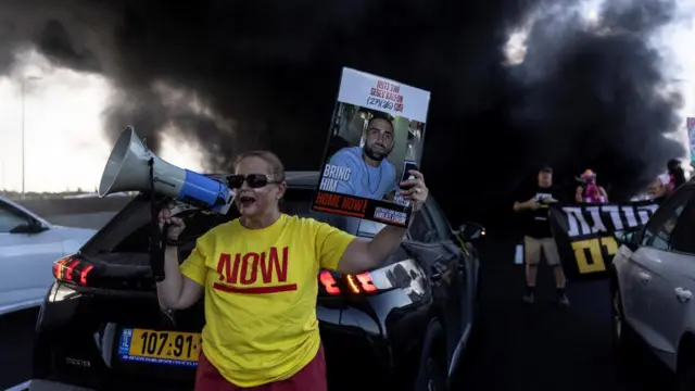 Protesters stand in front of cars on a main road with smoke in the background