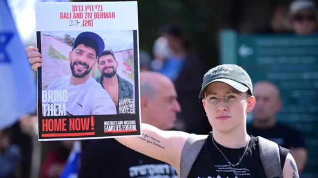 Adi holds up a poster with the images of two men - and a caption reading 'Bring them home now'