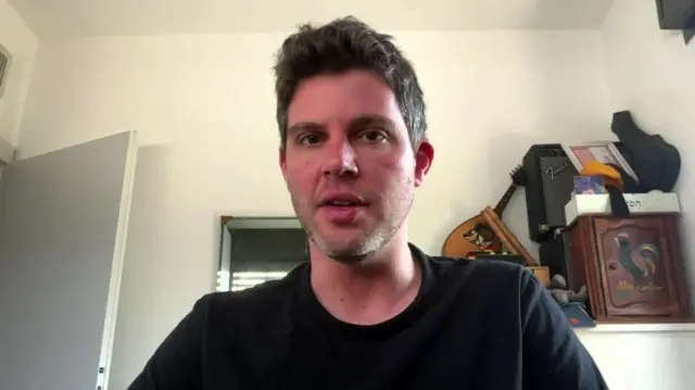 A man in a black long sleeve top looks at the camera, he's in a room with white walls and some musical instruments behind him