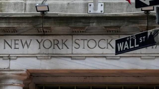 A building with letters engraved saying 'New York Stocks'. In the foreground, a street sign saying 'Wall Street'.