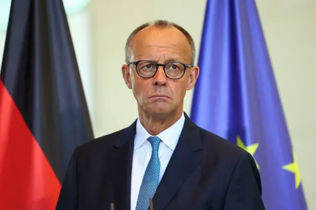 German Chancellor Friedrich Merz holds a press conference with Canadian Prime Minister Mark Carney (not pictured. Merz is wearing black spectacles and has a dark black suit with a turquoise patterned tie. He is standing in front of a German and EU flag with a serious expression on his face.