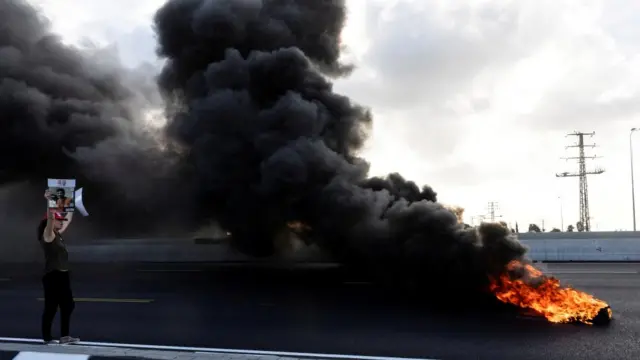 A woman holds posters in the middle of the road next to thick black smoke rising from a tyre fire