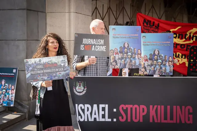 (Two people demonstrating at a protest in Brussels honouring fallen journalists in Gaza. One woman is holding a placard which reads "Israel stop killing Palestinian journalists in Gaza". She is standing next to a black banner with red and white writing which says "Israel Stop Killing". Next to here is a man holding a black placard which reads "Journalism is not a crime" in red and white capital letters. There are two placards of journalists who have been killed in Gaza being held by someone we can't see. Behind them is a red banner for the National Union of Journalists)