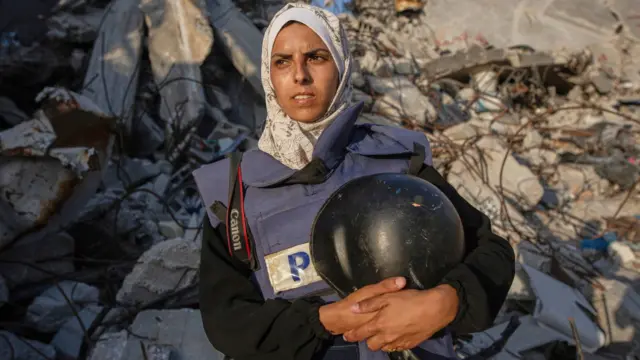Mariam Dagga holds her press helmet and wears a headscarf. She stands in front of rubble
