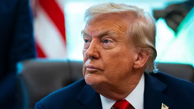 Donald Trump in blue suit with red tie. Looking to the side. Sat in a black chair.