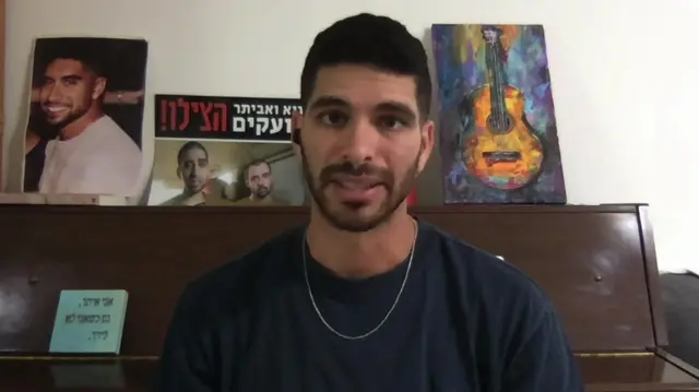 A man with a navy blue t shirt on and a chain sits in front of a wooden table, behind him are posters with his brother on them