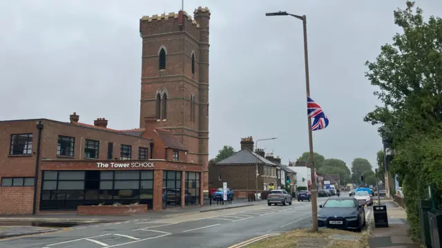 Epping High Street, as pictured from the side of the road. Cars are being driven along it on a grey day, while other vehicles are parked by the kerb. There is a church with a tall spire and shops lining the road.
