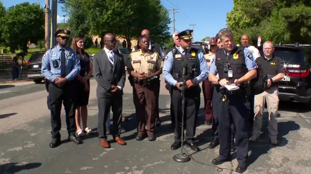 A group of police officers and other officials stand on a pavement