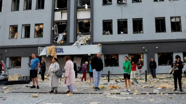 The British Council building, which is at least five storeys tall, and all of its windows are smashed out. A small group of people, some in their pajamas, are standing out the front looking at the damage. There is debris all over the ground out the front.
