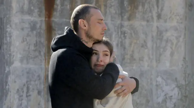 A man in a black hoodie keeps his eyes closed as he embraces a crying woman wearing a white hoodie, a damaged grey wall behind them