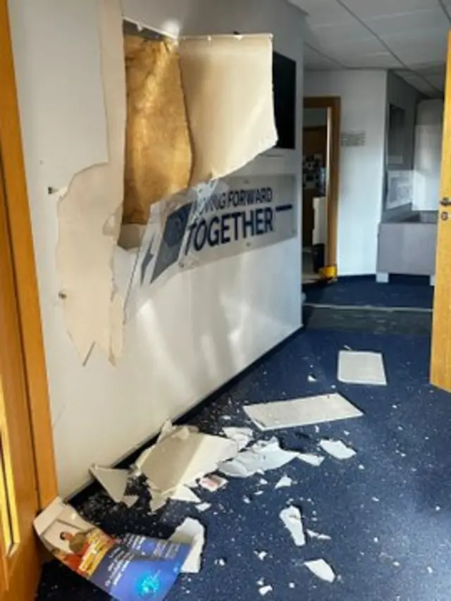 A sign on the inside of an office wall that says "moving forward together" is bent and damaged, with parts of the wall lying on the floor from the strike blasts.