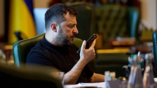 Volodymyr Zelensky holds up an iPhone as he speaks into it from behind a desk.