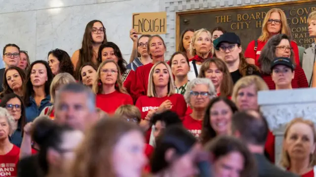 A hand holds a placard reading "enough" among a crowd listening to a speech