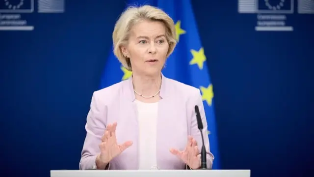 Ursula von der Leyen in white shirt and pick blazer, EU flag with blue background and yellow stars behind her.