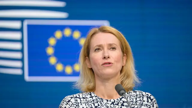 EU High Representative for Foreign Affairs Kaja Kallas in a black and white dress speaks into a thin black mic in front of a blue background bearing a EU flag