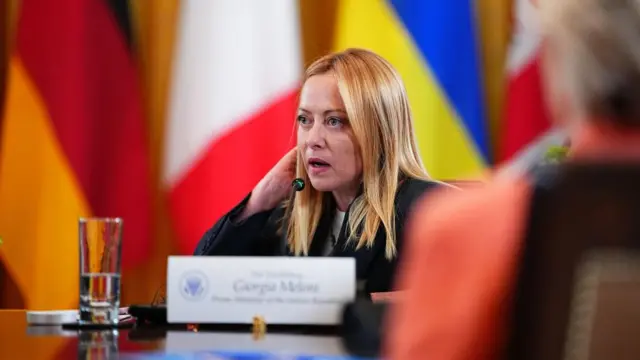 Giorgia Meloni speaks into a microphone from her seat at a table.