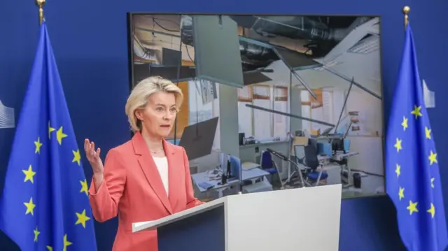 Ursula von der Leyen in a coral pink blazer and white shirt gestures as she speaks from a lectern flanked by EU flags. A picture of the inside of a room damaged by air strikes is shown on a screen behind her