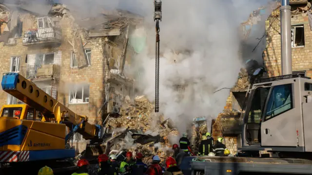 Rescue teams operating outside a bombed out residential building in Kyiv, grey smoke rising from a collapsed part of the five-storey building, the top floor entirely caved in