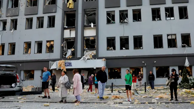 A large group of people stands in sleep wear on the street outside a building in central Kyiv damaged by a missile strike. Debris, damaged windows and insulation are strewn along the street, the building bears the logo of the British Council