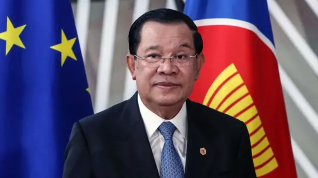 Hun Sen in a black suit and light blue tie, standing in front of the European and Asean flags.