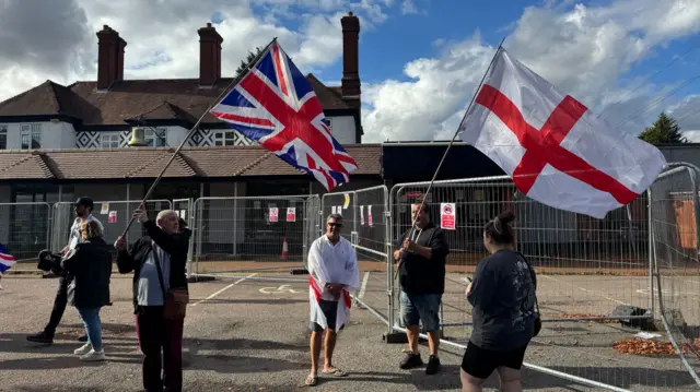 Six people. One holding a Union jack flag, another the St George's flag outside the hotel.