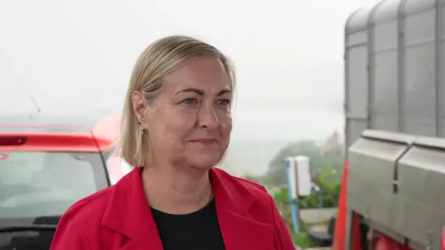 Alison Hume, wearing a red coat over a black top, stands alongside vehicles.