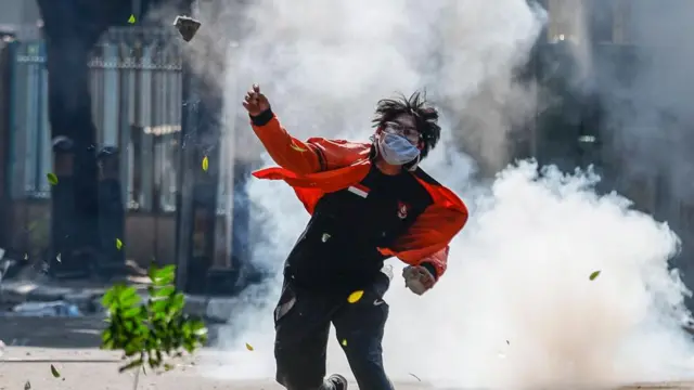 Protestor wearing an orange jacket and face mask throws a rock with tear gas smoke behind him