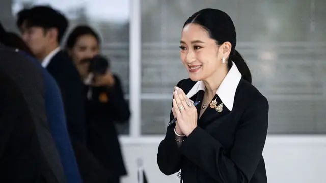 Paetongtarn smiling with her palms pressed together. She is wearing a black suit and sporting a sleek ponytail.