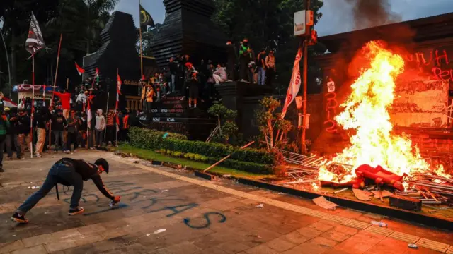 A protester sprays paint on the sidewalk near burning road barriers set on fire during a protest outside Jakarta's police headquarters