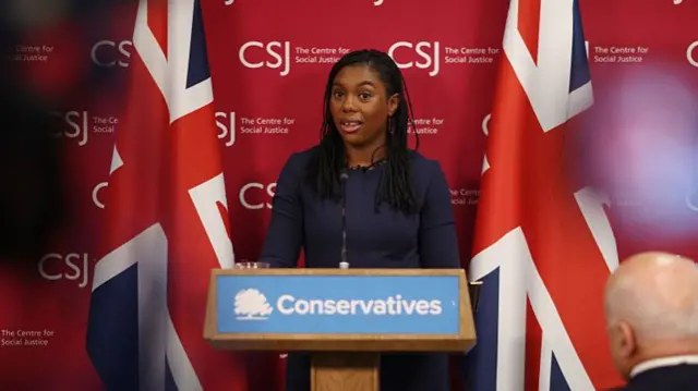Conservative leader Kemi Badenoch stood behind a podium that reads 'Conservatives'. UK flags behind her.