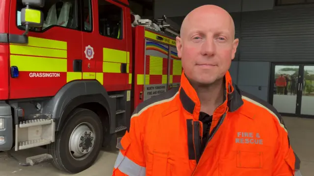 Jonathan Dyson, wearing an orange hi-vis jacket, stands alongside a fire engine