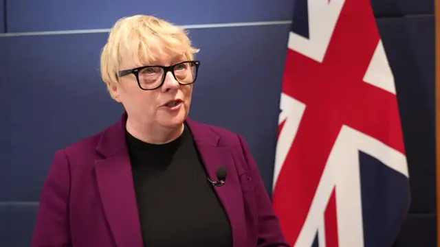 A woman in a purple jacket and black jumper stands in front of a union jack