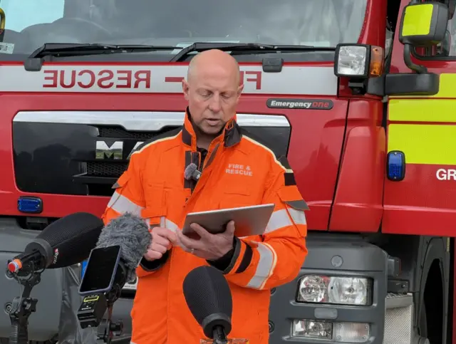 Jonathan Dyson with bald head wearing an orange jacket that says "fire and rescue" on it. He is stood in front of a fire engine and is speaking from a tablet device in front of three microphones.