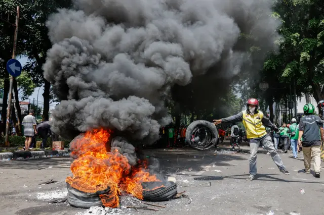Motorcycle driver throws a tyre onto a pile of burning tyres