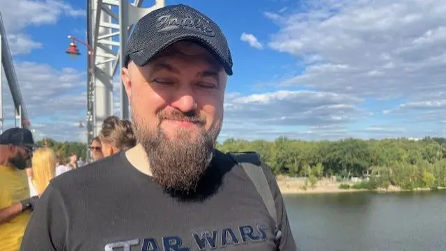Oleksandr Zvezdogliad in a grey Star Wars t-shirt, with dark cap and eyes closed. He stands on a bridge with water behind him and trees.