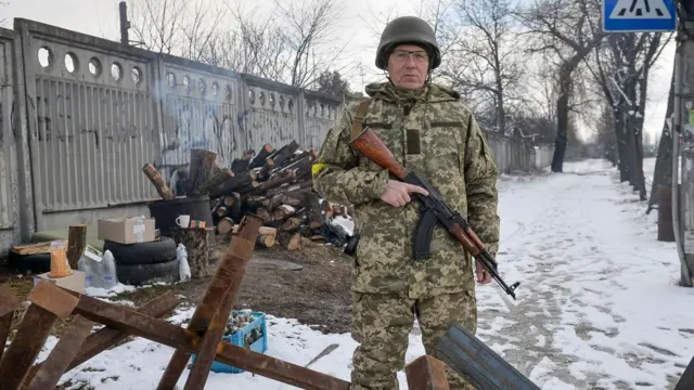 Andriy Parubiy stood outside with snow and trees behind him, logs to his left. He is in military uniform holding a rifle, looking at the camera.
