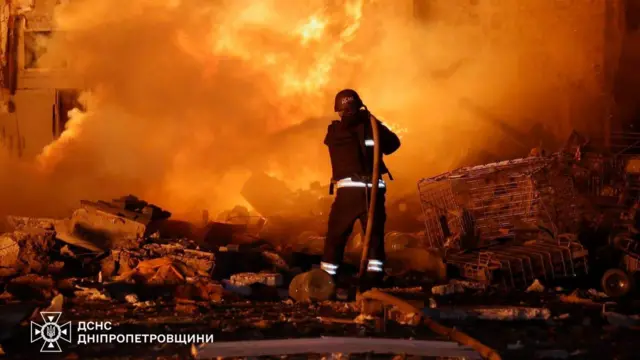 An emergency service worker holds a hose and seeks to extinguish a fire. They have their back to the camera and rubble covers the floor