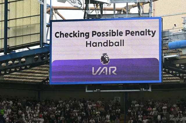 The big screen displays the news that a VAR (Video Assistant Referee) review is looking at a potential handball in the panalty area