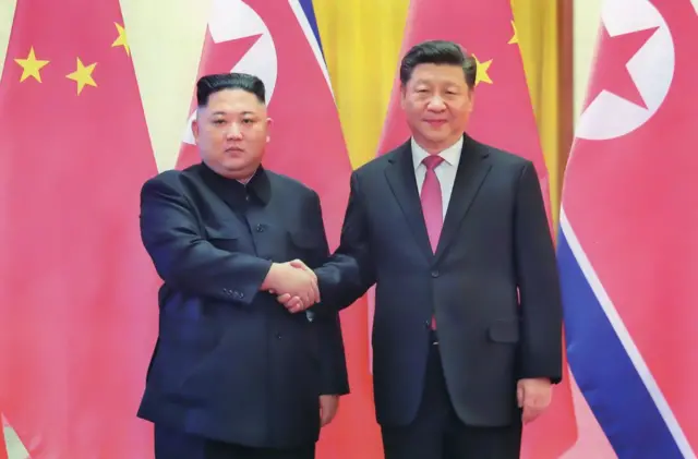 China's President Xi Jinping shakes hands with North Korean leader Kim Jong-un. They both wear black suits and have neutral expressions.