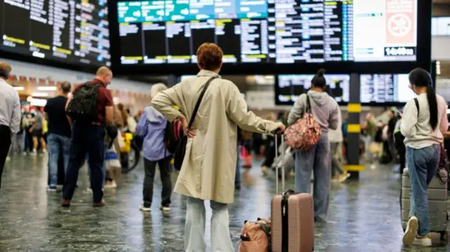 A woman in a beige trench coat (C) observes the travel information board inside a train station, other passengers with bags waiting around her